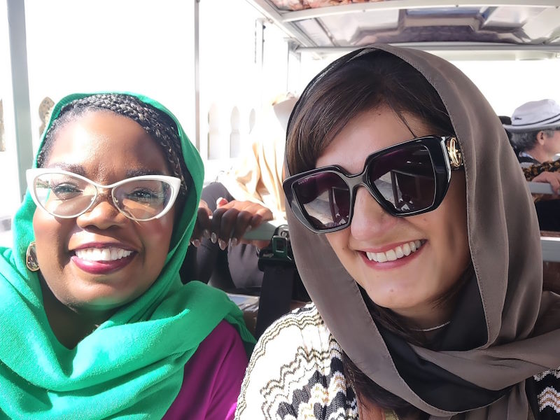 Two smiling women wear head coverings in a vehicle.
