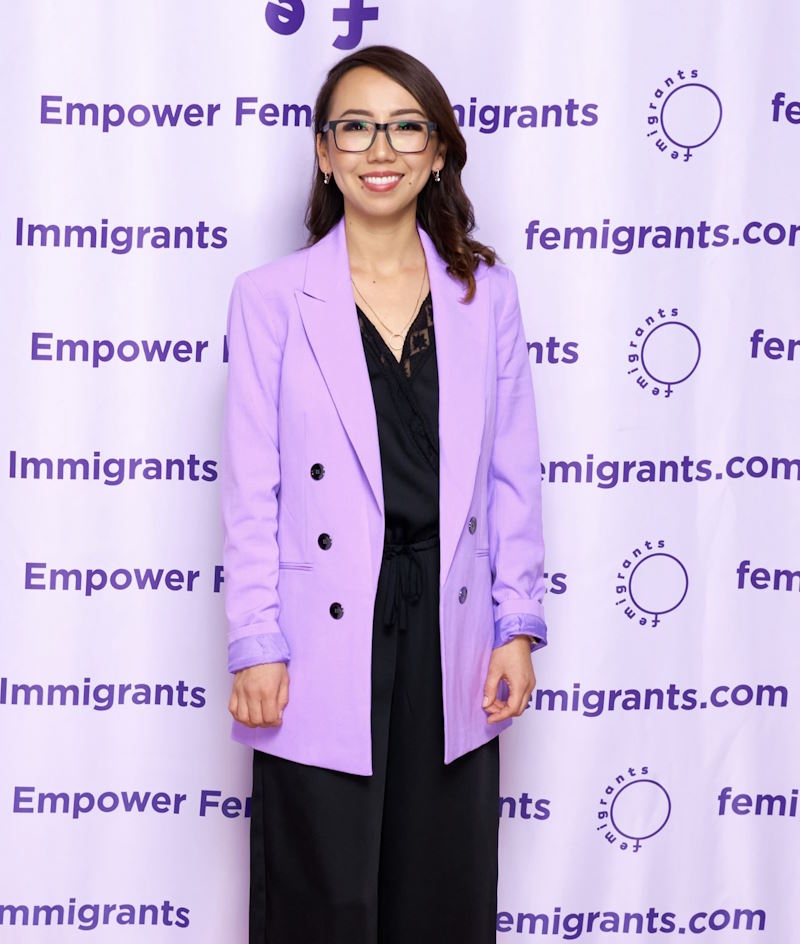 Kani smiling in front of a step and repeat banner, wearing glasses and a lilac coat.