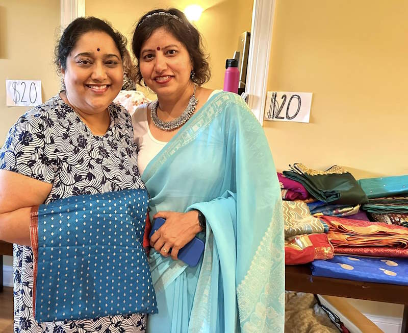 Two smiling women next to a stack of colorful sarees.