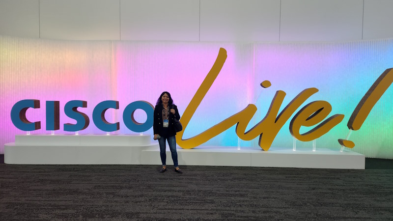 Nayeli stands in front of a large blue and yellow Cisco Live logo.
