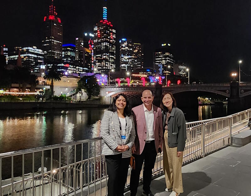 Nayeli, Dave, and Esther stand with downtown Melbourne’s skyline lit up at night.