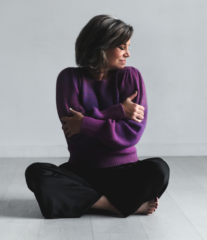 Rana wears a purple sweater and sits cross-legged, giving herself a hug.