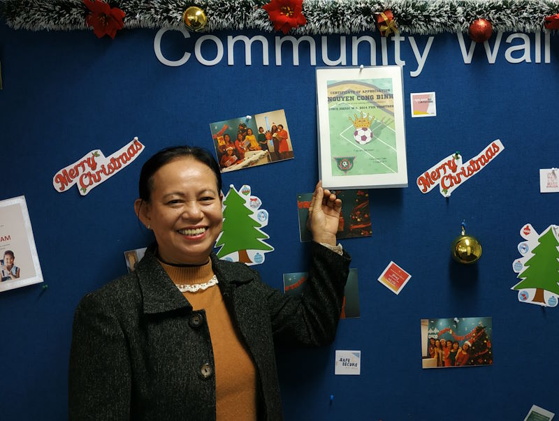 Binh’s mom smiles with a certificate on a colorful community wall.