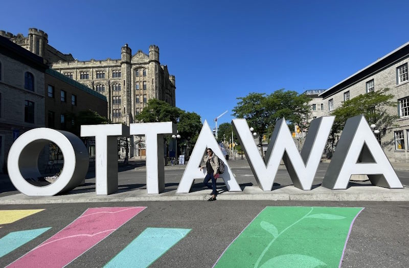 Madhu stands in front of large white letters that  spell Ottawa.