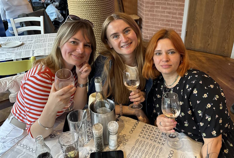 A group of three women holding wine glasses sit at a table.