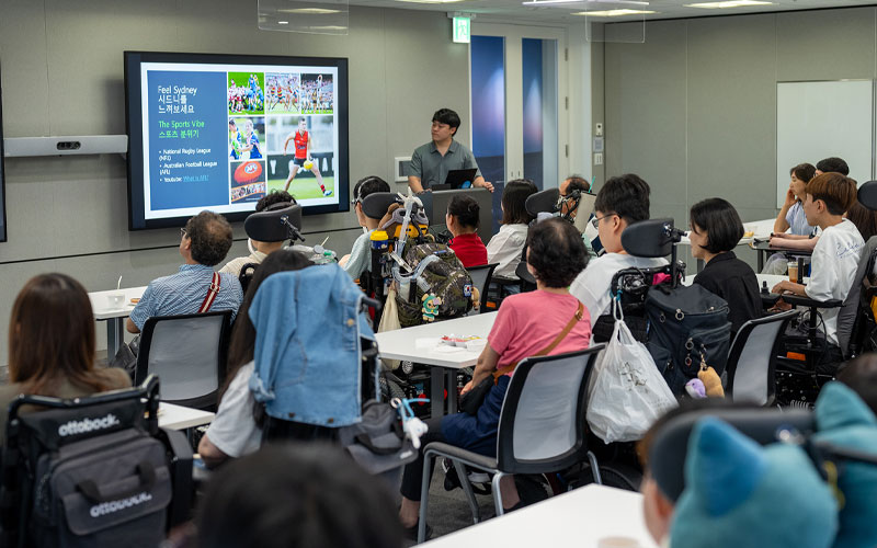 A man in a gray shirt stands in front of the room, presenting a slide with sports images to the participants.