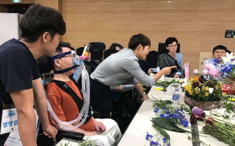 Five people, one in an oxygen mask, at a table filled with flowers to make flower arrangements.