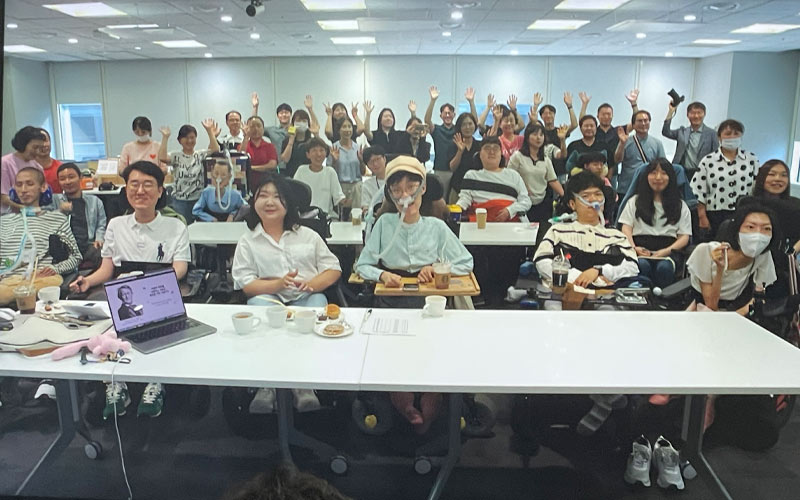 Many people, some in wheelchairs and oxygen masks, sit at tables at a Cisco office, while those in the back wave. 