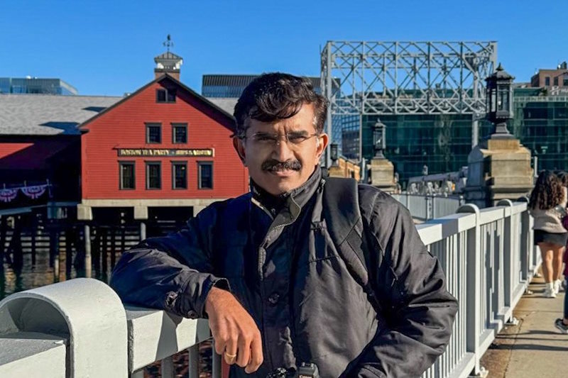 Rakesh stands on a bridge in front of a red building in Boston. 