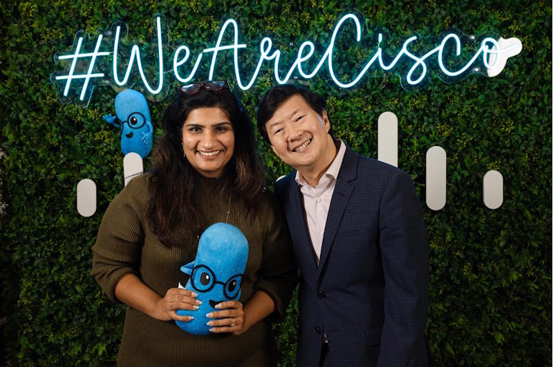 Two people smiling in front of a green plant background.