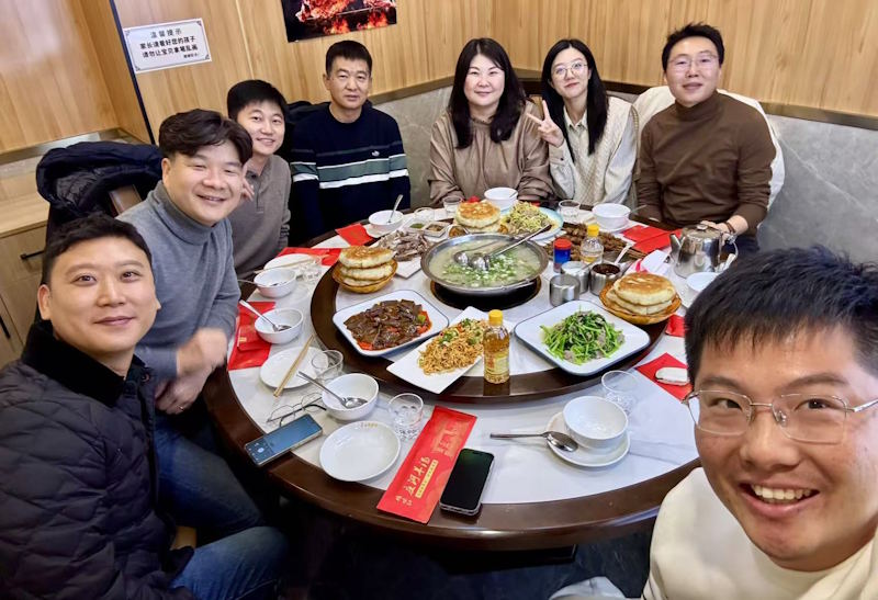 Smiling colleagues sit around a circular table, sharing food.