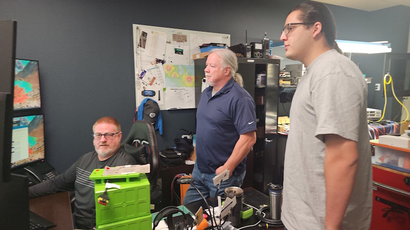 Three people look at a computer screen with technical equipment in the background.