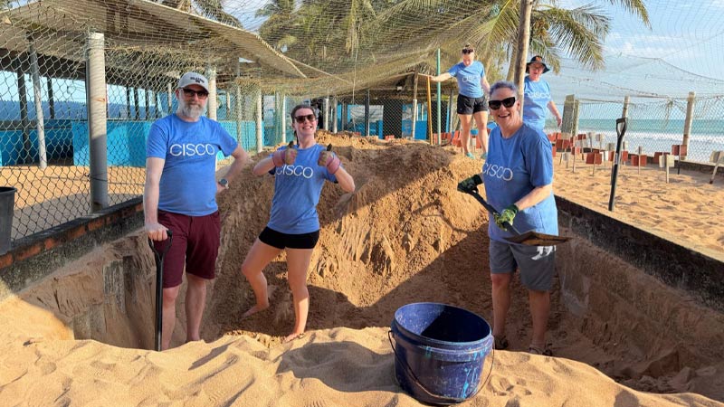 Five people wearing blue Cisco t-shirts stand in a large sand pit, digging with a spade.