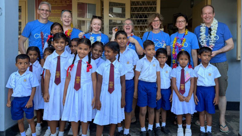 A group of school children in uniforms stands in front of adults wearing blue t-shirts and flower garlands.