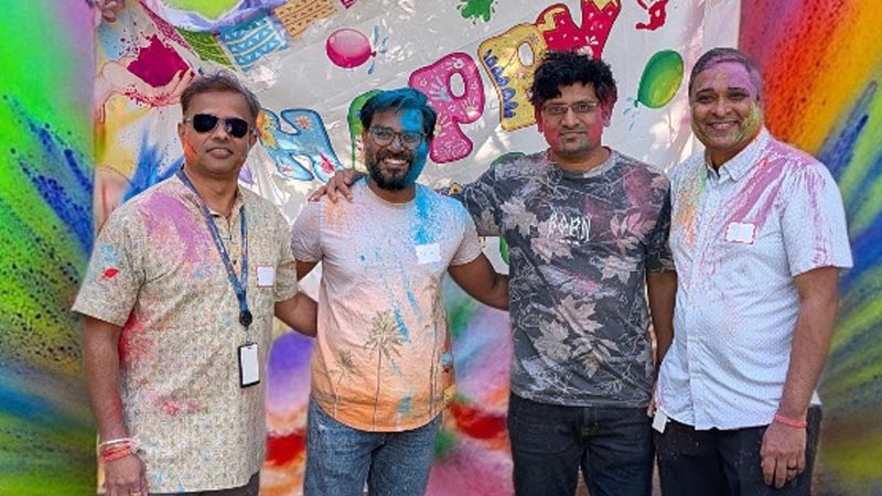 Four men covered in colorful powder stand in front of a vibrant, multicolored backdrop.