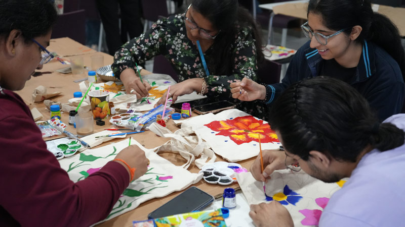 People seated around a table paint fabric bags with floral designs. 