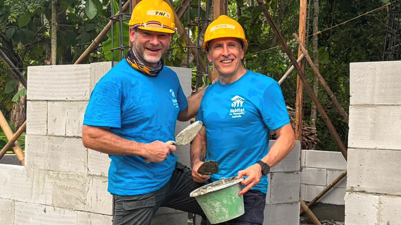 Two people wearing yellow hard hats and blue volunteer t-shirts hold a bucket of cement and a trowel.