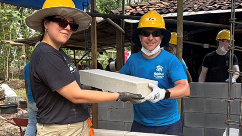 Two people wearing yellow hard hats pass a brick to each other.