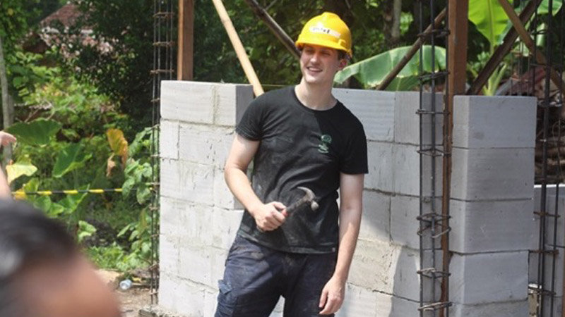 A person stands in front of a half-built brick wall, wearing a yellow hard hat and holding a hammer.