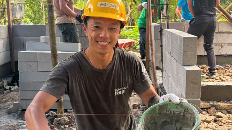 A person wearing a yellow hard hat holds a bucket of cement and a trowel.