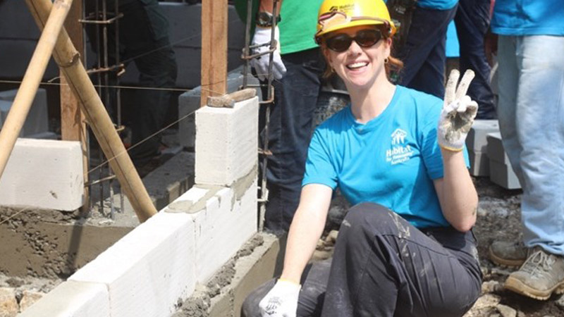 A person wearing a yellow hard hat and sunglasses holds up a peace sign, kneeling in front of a half-built wall.