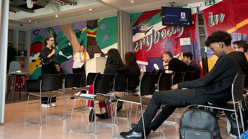 A woman presents to an audience at an indoor event space with a colorful mural in the background.