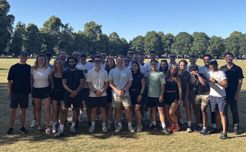 A group of people stands on grass in a park with trees and clear blue sky.